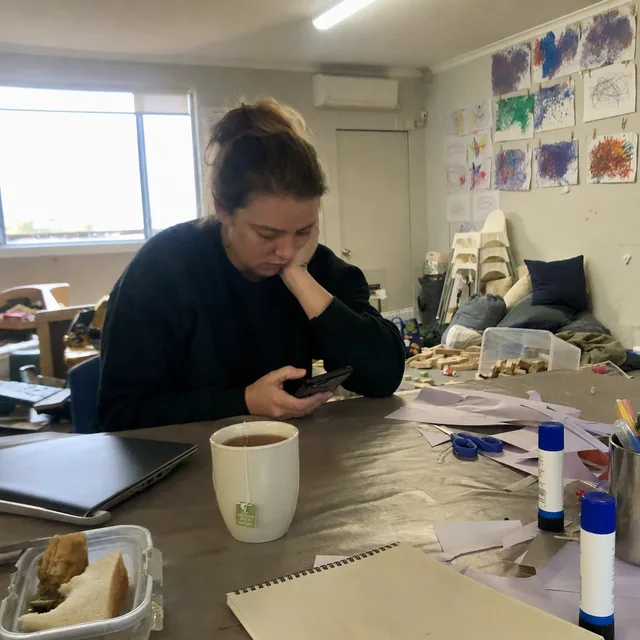 An educator sitting at a cluttered staff table late in the day, editing photos on her phone