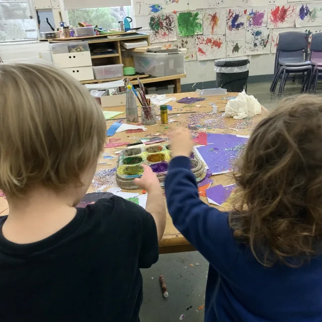 Two children seen from behind at a craft table covered in paint, paper and glitter